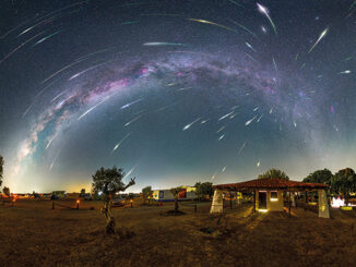 A stunning night sky composite photograph shows dozens of bright Perseid meteors streaking across the sky above a rural landscape, with the Milky Way arching overhead. The foreground features trees, a house, and softly lit structures, creating a serene countryside scene.