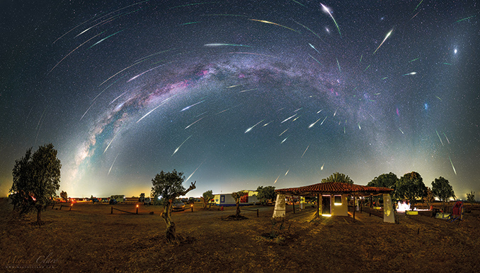 A stunning night sky composite photograph shows dozens of bright Perseid meteors streaking across the sky above a rural landscape, with the Milky Way arching overhead. The foreground features trees, a house, and softly lit structures, creating a serene countryside scene.
