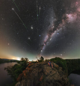 A panoramic night sky photo shows a person sitting on a rocky outcrop overlooking a river and forest, with the Milky Way stretching across the sky and numerous meteors streaking across the sky.