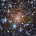 A dense star cluster filled with thousands of orange and blue stars against a dark sky. The bright blue stars create a sparkling effect around the cluster’s glowing core.